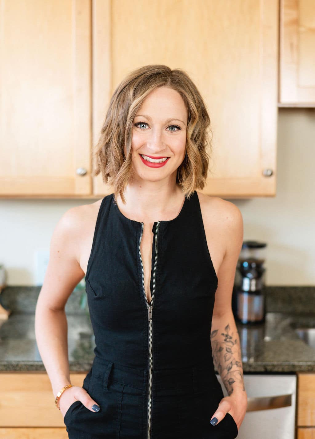 Kaytee, a white woman with short blonde hair, wears a black jumper in a kitchen, smiling